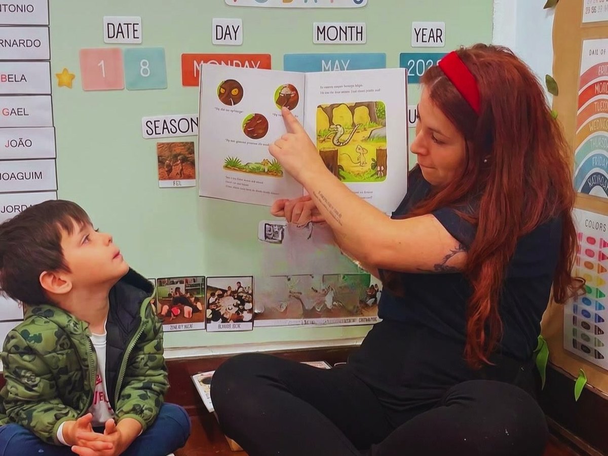 Professora mostrando um livro a uma criança diante do quadro da rotina em inglês